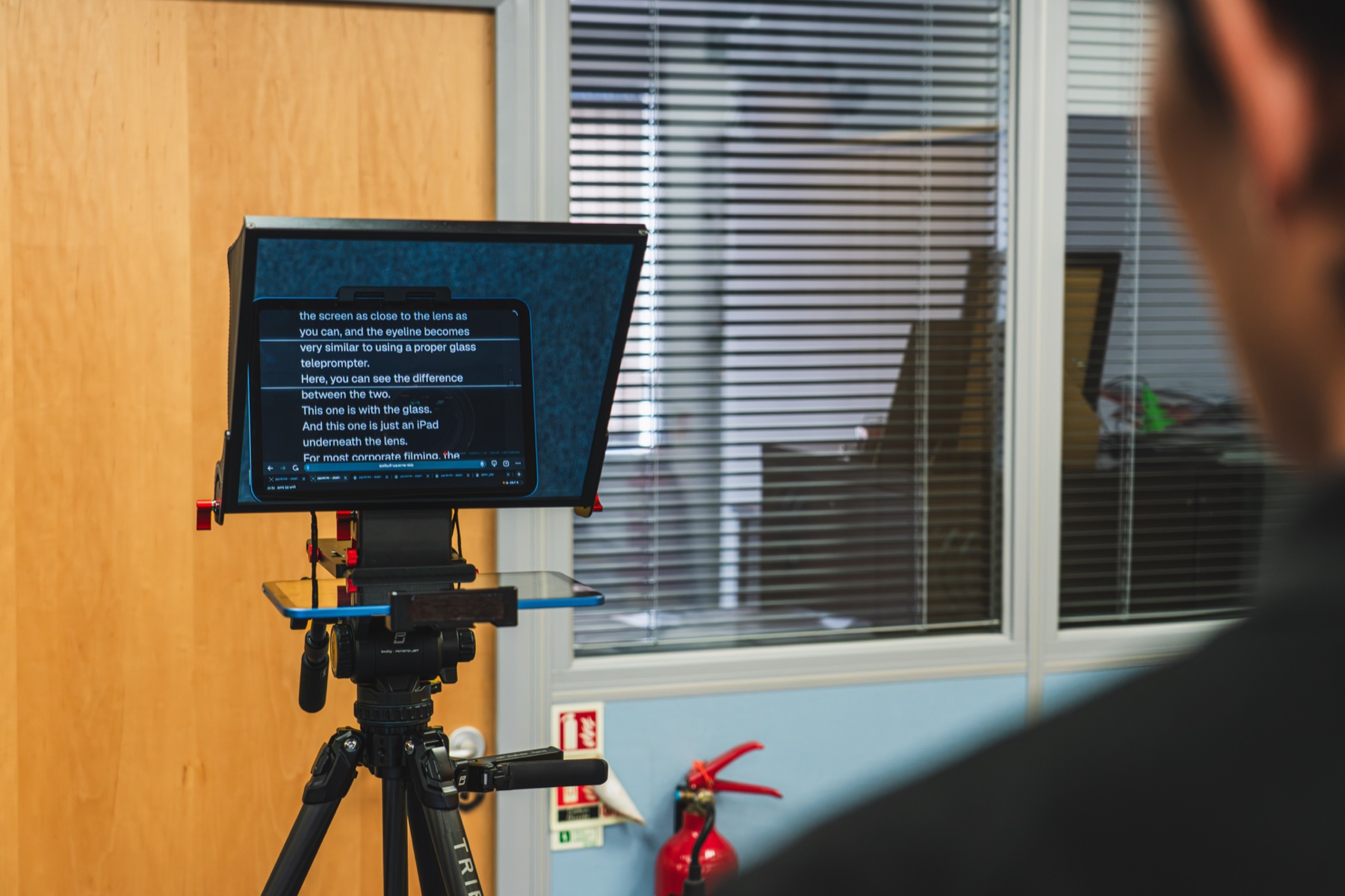 Over the presenter's shoulder mid-read, with the script floating on the angled glass directly in front of the lens.