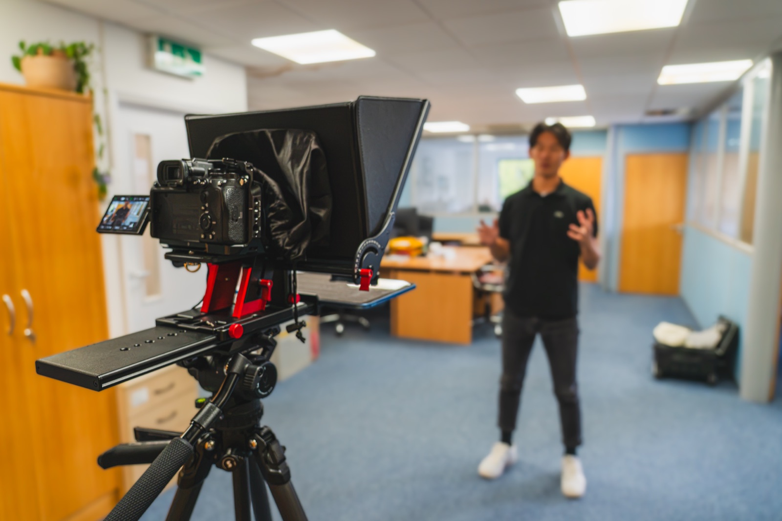 A filming day at the studio: a presenter stands on his mark while a cinema camera and teleprompter rig face him from a tripod in the foreground.